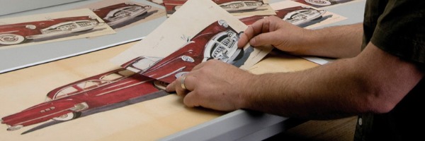 Close up on a printed image of a red car lain out on a table. A conservator's hands can be seen, holding a digitally reproduced image of the car up against the original. 