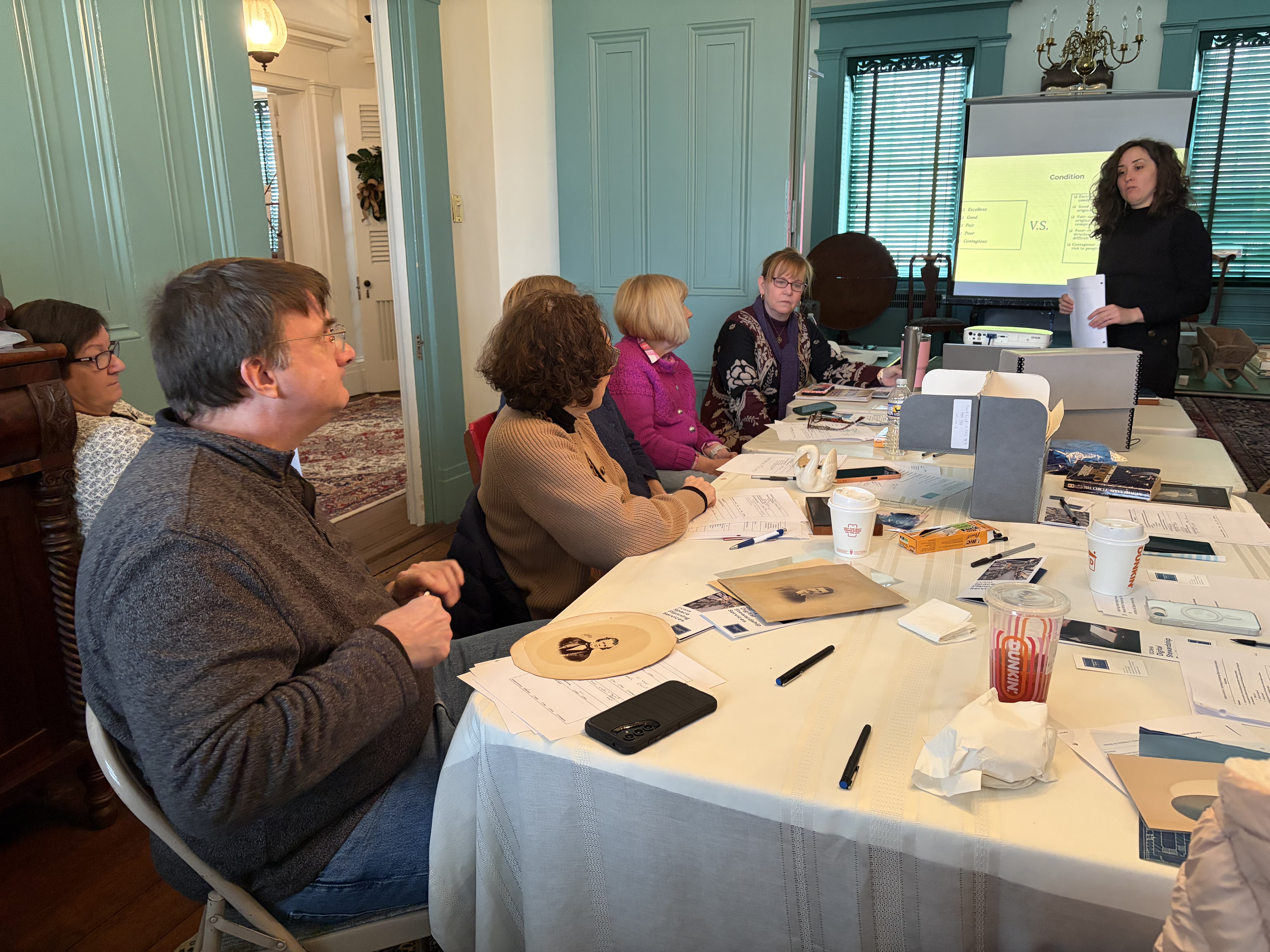 A group of participants seated at a long table during a preservation workshop. They are examining historical photographs and documents while a presenter stands at the front next to a projector screen labeled "Condition." The room features historic architectural details and teal-painted walls.