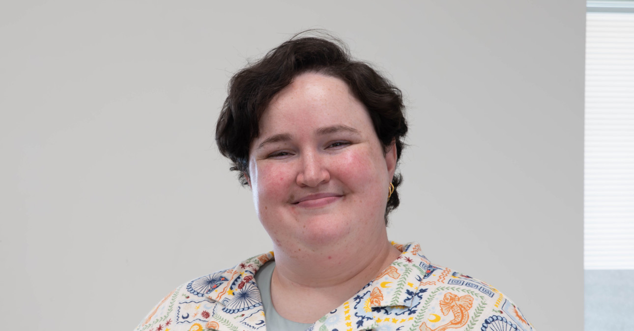 Manager of Digital Imaging, Gray Ortega, stands in front of a white background, smiling. They are wearing a patterned shirt and have short brown hair.