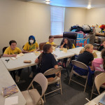 Group of young students sitting around folding tables engaged in a hands-on bookbinding activity with paper, scissors, and tools.