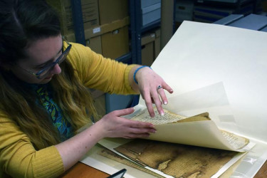 An archivist with glasses and a yellow cardigan carefully lifts a protective sheet to examine a fragile, weathered historical document in an archive room filled with storage boxes.